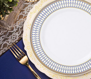 A beautifully set table with a decorative white porcelain plate featuring an intricate blue and gold design on the rim. The plate rests on a lace tablecloth with a navy blue placemat underneath. A gold fork is placed to the left of the plate, complementing the elegant setting.