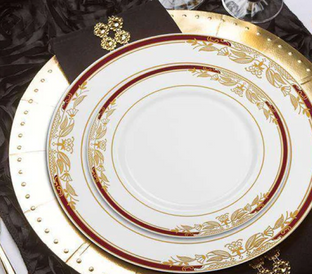 An elegant table setting featuring stacked, ornate plates with gold and red detailing, placed on a gold charger plate. The setup includes a black napkin with gold embellishments, all set on a black textured tablecloth, adding a touch of luxury.