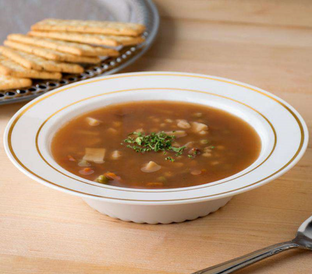 A bowl of hearty vegetable soup garnished with chopped herbs sits on a wooden table. Beside it, there is a silver tray with an arrangement of crackers, and a silver spoon rests nearby. The soup bowl is white with a decorative gold rim.