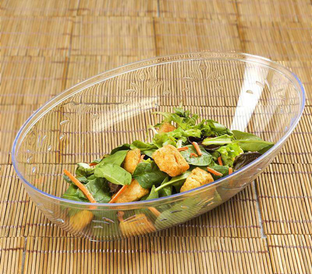 A translucent plastic bowl on a bamboo mat background contains a mixed green salad with leafy greens, carrot sticks, and croutons.