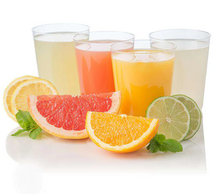 Four glasses of juice are lined up, each filled with a different colored drink: light yellow, pink, orange, and white. In front of the glasses are cut slices of lemon, grapefruit, orange, and lime, along with sprigs of fresh mint.