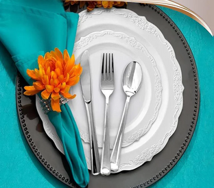 Table setting with a white decorative plate on a larger black plate, surrounded by a beaded edge. On the plate are a fork, knife, and spoon. A turquoise napkin with an orange flower is placed on the left side, all set on a turquoise tablecloth.