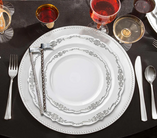 Elegant table setting featuring a white plate with ornate silver designs, silverware on either side, and two small decorative hammers wrapped in ribbon on the left. The setting includes a fork, knife, and spoon, with assorted glasses filled with colored liquids.