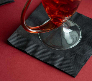 A glass of red liquid is placed on a folded black napkin, which rests on a red surface. The handle of the glass curves elegantly, and the napkin's edges have a striped texture.