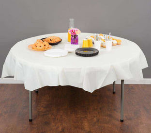 A round table with a white tablecloth is set with a variety of breakfast items, including donuts, pastries, cups of yogurt, and a pitcher of orange juice. A small vase with flowers is also placed at the center of the table. The background is a plain gray wall.
