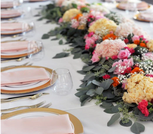 A beautifully set table decorated with a vibrant centerpiece of mixed flowers, including hydrangeas, roses, and baby's breath. Each place setting features a gold-rimmed plate, folded pink napkins, and a stemless wine glass, all on a white tablecloth.