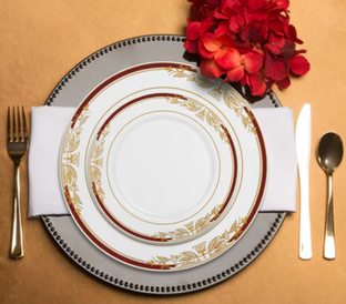 A table setting featuring a white plate with a gold and red border on a larger grey plate with a beaded rim. The setting includes a gold fork, knife, and spoon. A white napkin is placed under the plates, and red floral decor rests above the plates on a tan background.
