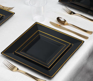 A table setting featuring square black plates with gold trim, stacked in three sizes. Gold-colored cutlery, including a fork on the left, and a knife and spoon on the right, are placed next to the plates. A clear plastic cup is visible in the background.