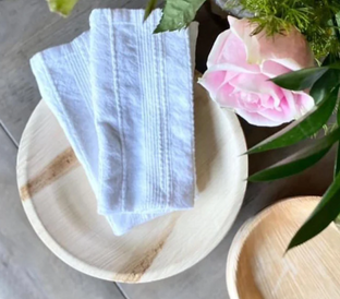 A white cloth napkin neatly folded on a wooden plate is beside a pink rose and some green foliage. Another empty wooden bowl is nearby. Both plates rest on a gray surface.