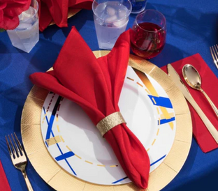 A place setting featuring a white plate with blue and gold accents, a gold charger plate underneath, and a red cloth napkin neatly folded into a decorative style with a gold napkin ring. The table also has gold cutlery, a water glass, and a glass of red liquid.