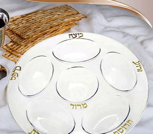 A Passover Seder plate with six clearly labeled sections in Hebrew, designed to hold symbolic foods for the Seder meal. Behind the plate, there are pieces of matzah (unleavened bread) and a white fabric.