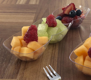 Three small, clear plastic bowls filled with fruit sit on a wooden table. The nearest bowl contains cantaloupe chunks and a raspberry. The middle bowl holds honeydew pieces topped with raspberries. The far bowl has mixed berries. A silver fork lies in front.