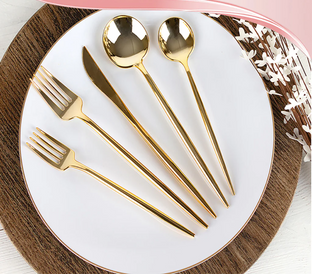 A round white plate with a rose gold rim features an elegant gold-colored cutlery set, including two forks, a knife, and two spoons, artistically arranged. The plate rests on a wooden placemat, and a pink ribbon and white flowers adorn the top corner.