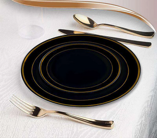A place setting on a white tablecloth, featuring nested black plates with gold rims, and gold-colored cutlery including a fork, knife, and spoon. A clear drinking glass is partially visible at the top left corner.