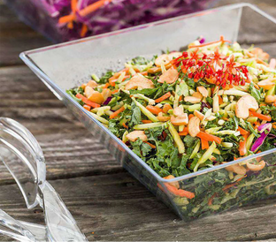 A clear square bowl filled with a colorful salad placed on a wooden surface. The salad includes chopped kale, carrots, bell peppers, cashew nuts, and garnished with small red flowers. Transparent salad servers are on the left side of the bowl.