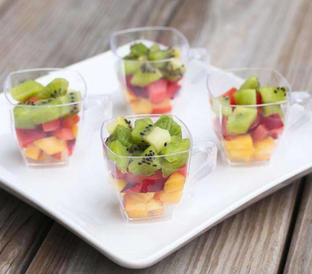 Four small, clear plastic cups filled with a colorful fruit salad consisting of chopped kiwi, red and yellow fruits are placed on a white square plate. The plate is set on a rustic wooden surface.