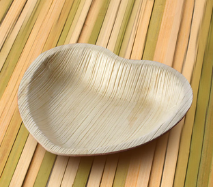A bowl made from biodegradable material shaped like a leaf sits on a wooden table with planks in various shades of beige, light brown, and green. The bowl has a natural, textured appearance with visible veins resembling those of a leaf.