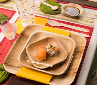 A table setting with eco-friendly wooden plates and cutlery, showcasing a piece of sushi with salmon and a sushi roll on the smallest plate. The table is adorned with colorful placemats, a glass of water, a small dish of soy sauce, and a cup.