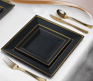 A set of square black plates with gold trim are neatly arranged on a white tablecloth. The set includes a large, medium, and small plate. Gold utensils—a knife, fork, spoon—are placed beside the plates, creating an elegant and sophisticated dining setup.
