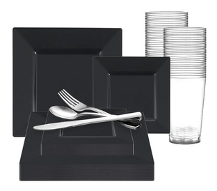 A set of black square plates and bowls of various sizes are arranged with stainless steel cutlery (knife, fork, and spoon) on top. Clear plastic drinking glasses are stacked in rows behind the plates.