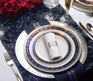 A formal table setting features an ornate navy blue and gold plate with a matching salad plate and a white napkin wrapped in a silver ring. Silver cutlery and two elegant glasses are placed on a dark floral table runner with red flowers in the background.
