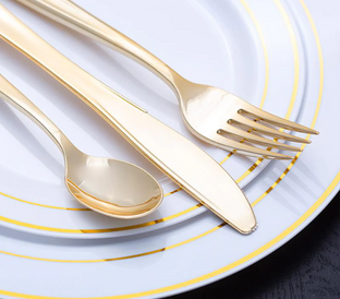 A gold spoon, knife, and fork are arranged on a white plate with gold accents. The utensils have a sleek, modern design, and the plate has concentric gold rings near the edges. The background shows a dark surface underneath the plate.