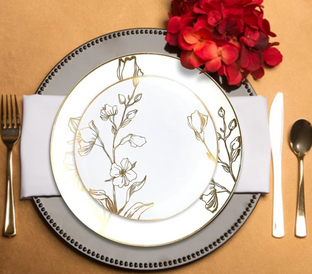 A formal place setting featuring a dinner plate and salad plate with floral designs, placed on a white napkin atop a gray charger adorned with a stitched edge. Silverware is arranged on either side, and a cluster of red flowers decorates the top right.