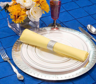 A table setting featuring a silver-edged white plate with a neatly rolled yellow napkin adorned with a silver napkin ring. The plate rests on a vibrant blue tablecloth. Nearby are a fork, knife, spoon, a glass of wine, and a vase with yellow and white flowers.