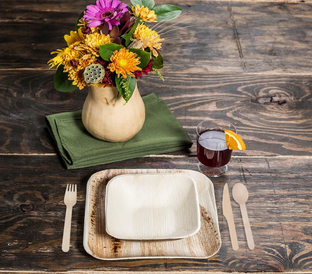 A wooden table set with eco-friendly square plates, a wooden fork and spoon, a green cloth napkin, a glass of juice with an orange slice, and a vase filled with colorful flowers. The natural textures and colors create a rustic and charming dining setting.