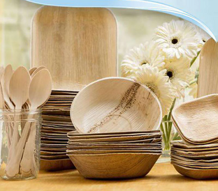 A collection of eco-friendly wooden tableware including plates, bowls, and spoons is neatly arranged on a table with a light orange tablecloth. A glass jar holding wooden spoons and a bouquet of white flowers are also placed together, adding a natural touch.