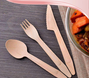A set of wooden cutlery including a fork, knife, and spoon is laid out on a dark wooden surface next to a glass bowl of cooked vegetables. The bowl contains carrots and green beans. A beige cloth is partially visible under the bowl.