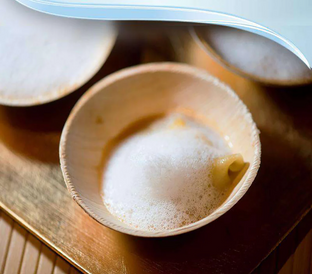 A close-up of a small wooden bowl filled with a foamy dish, featuring a piece of pasta partially submerged in the froth. The bowl rests on a wooden surface, with another similar bowl partially visible in the background.
