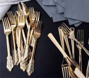 A collection of gold-colored flatware, including ornate and plain forks, knives, and spoons, arranged on a dark surface. Some pieces have intricate designs on their handles, while others are smooth. Gray napkins are visible in the background.