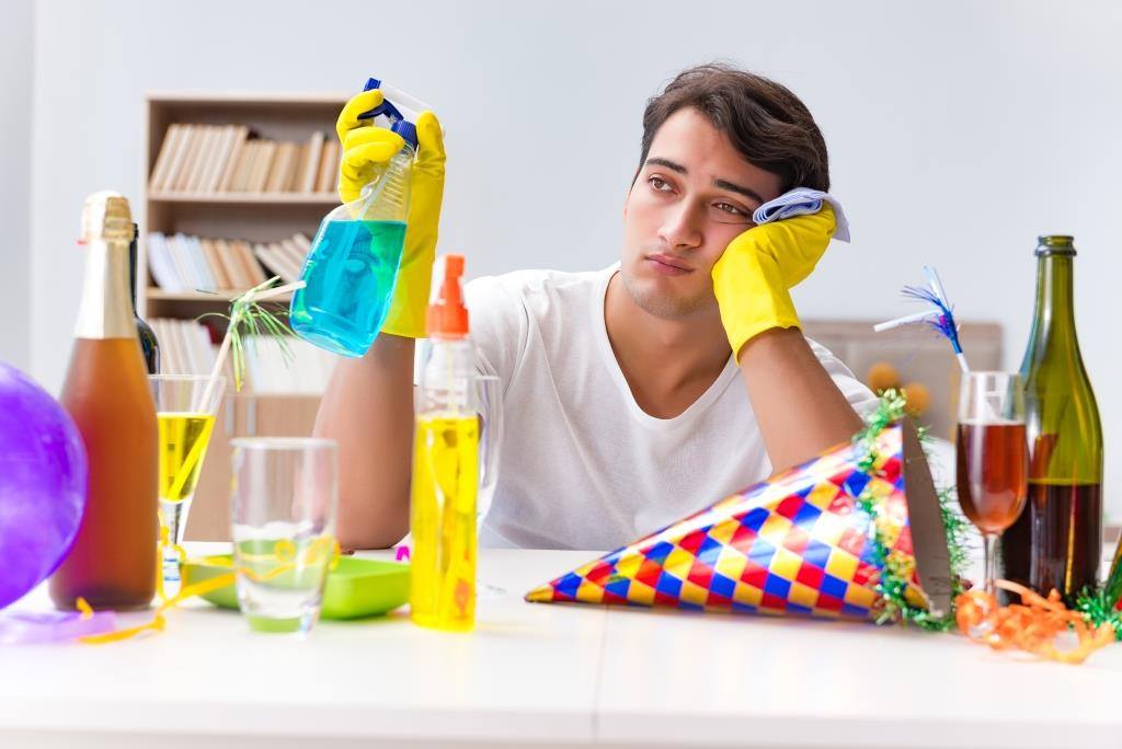 A man sits at a table surrounded by balloons, bottles, and dirty dishes, reflecting on the choice between using paper plates vs. washing dishes.