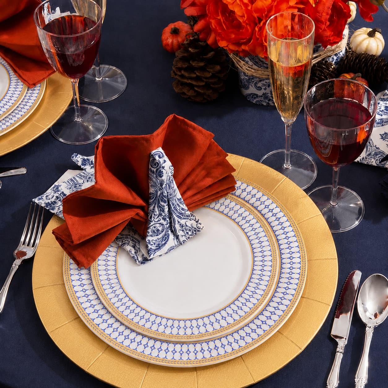 Elegant Thanksgiving table setting with blue and gold disposable plates, gold charger, rust napkin, clear stemware, and autumn floral decor.