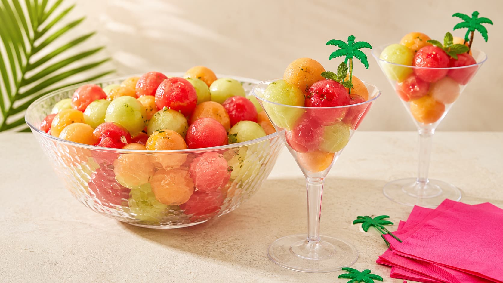 Drunken melon balls served in a clear plastic bowl with plastic martini glasses, pink cocktail napkins, and palm picks for a summer party