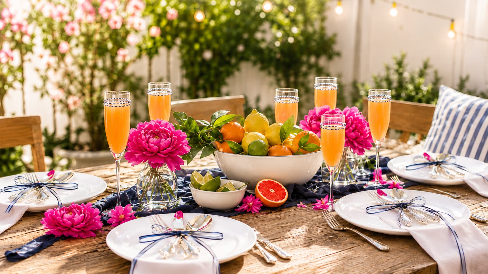 Outdoor summer party table with white disposable plates, citrus centerpiece, pink flowers, and orange drinks