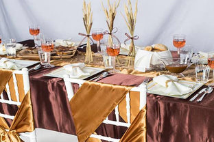Elegant table setting with white chairs adorned with brown and gold sashes. The table features brown tablecloths, white plates, silver cutlery, glasses of rosé wine, and rolled napkins. Wheat decorations and bread rolls add a rustic touch to the arrangement.