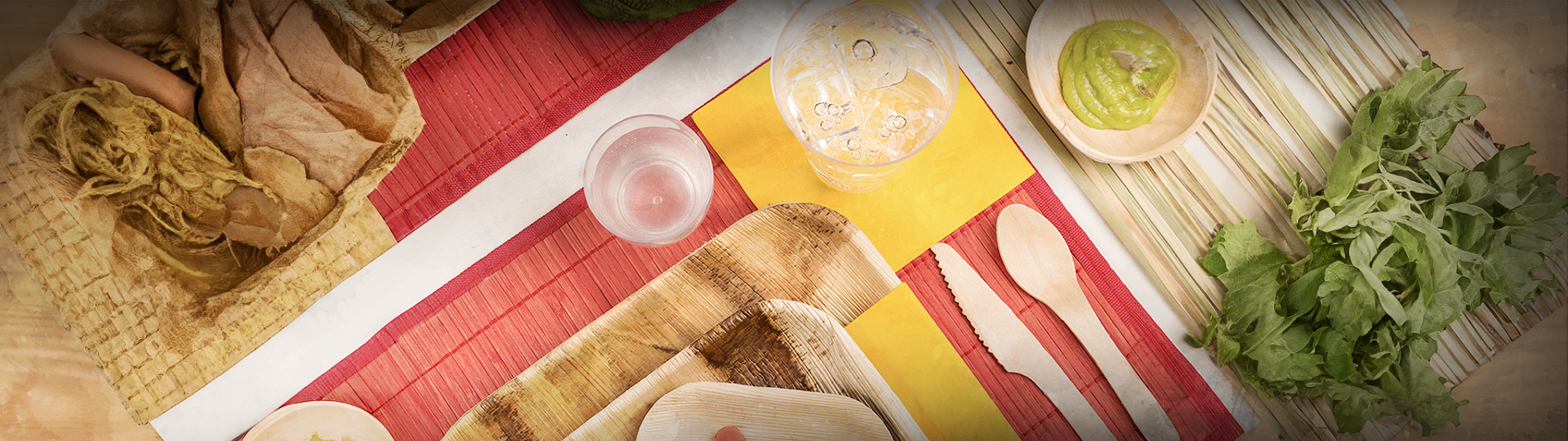 A variety of food items and utensils are laid out on a tabletop with colorful placemats. The items include bread, a glass of water, a wooden spoon and fork, a bowl of green dip, and fresh greens. The scene captures a vibrant and inviting meal setup.