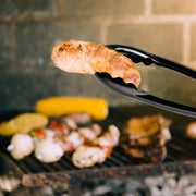 Black heavyweight PP serving tongs resting on a buffet platter, hygienic disposable serveware for weddings, receptions, and holiday parties.
