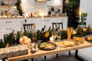 A festive dining table is set with plates of salad, glasses of champagne, a bottle of wine, breadsticks, and various other dishes. The table is decorated with a garland of greenery, candles, and holiday ornaments. A wrapped gift sits on the edge of the table.